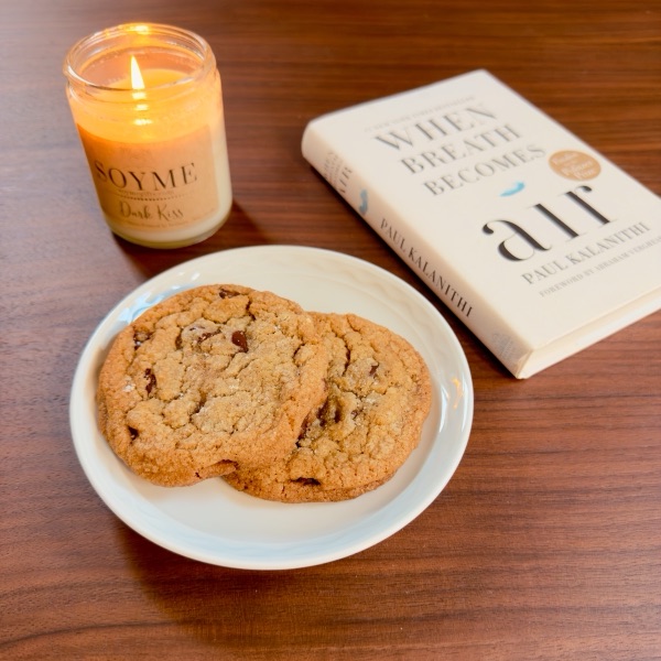 Two brown butter chocolate chip cookies on a white plate, next to a lit candle and a book on a wooden table